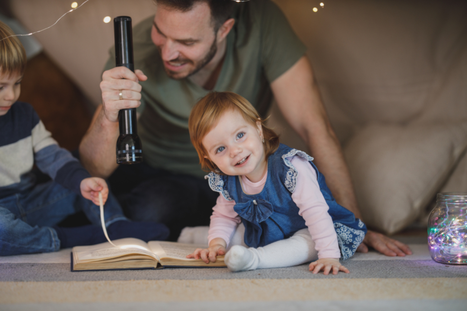 Une famille lit sous un tipi de couvertures, à la lampe torche